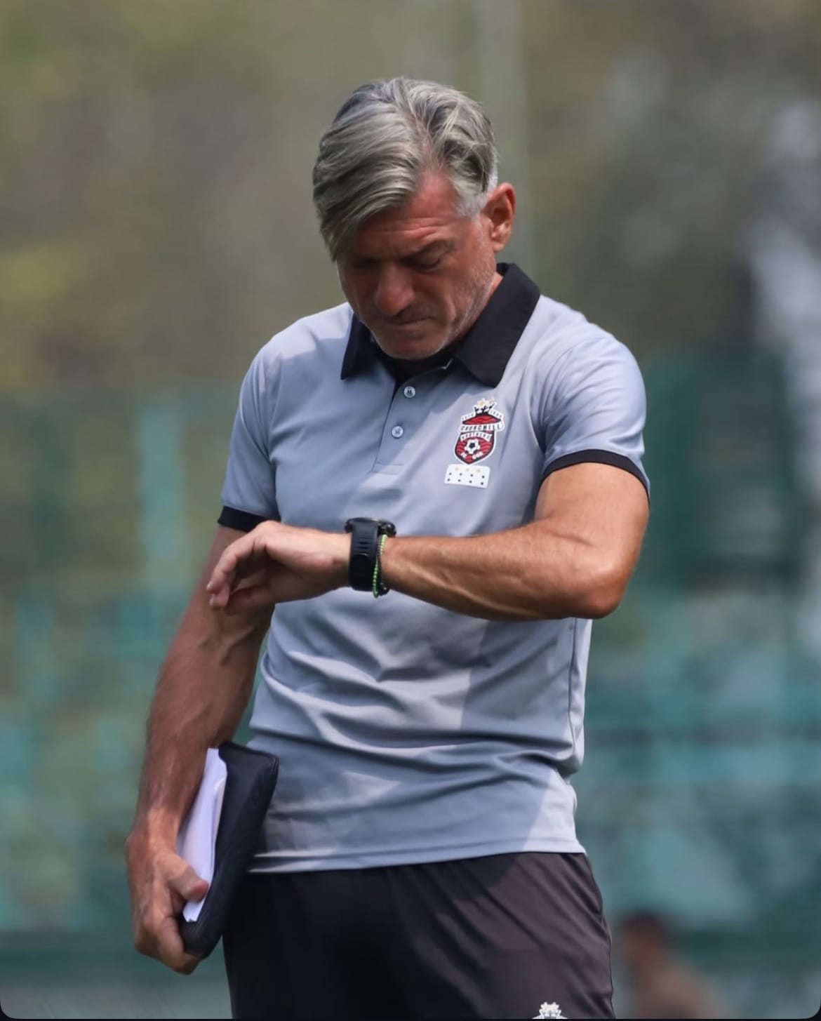 Coach wiping his arm looking at his watch, holding a notebook.