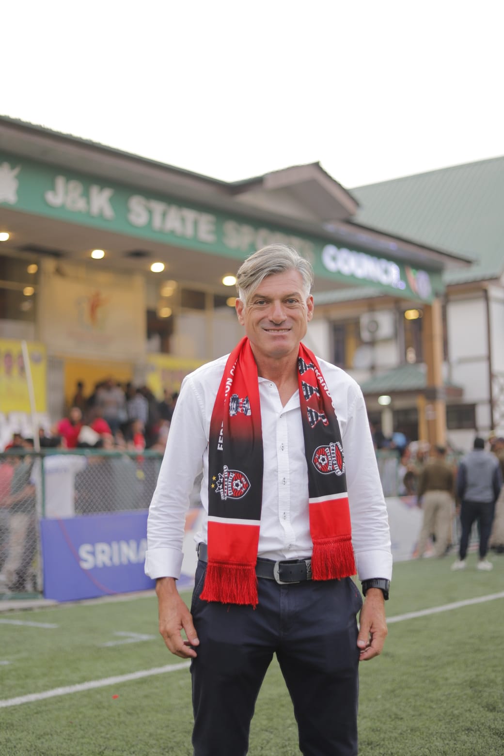 Man standing on a sports field, smiling at the camera, wearing a scarf. A man with short and grey-silver colored hair is s...